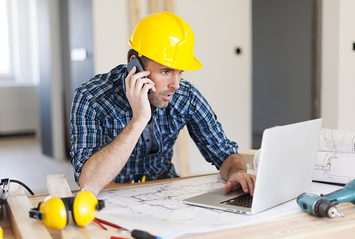 Contractor in a hard hat works on his website on a laptop while talking on a smartphone